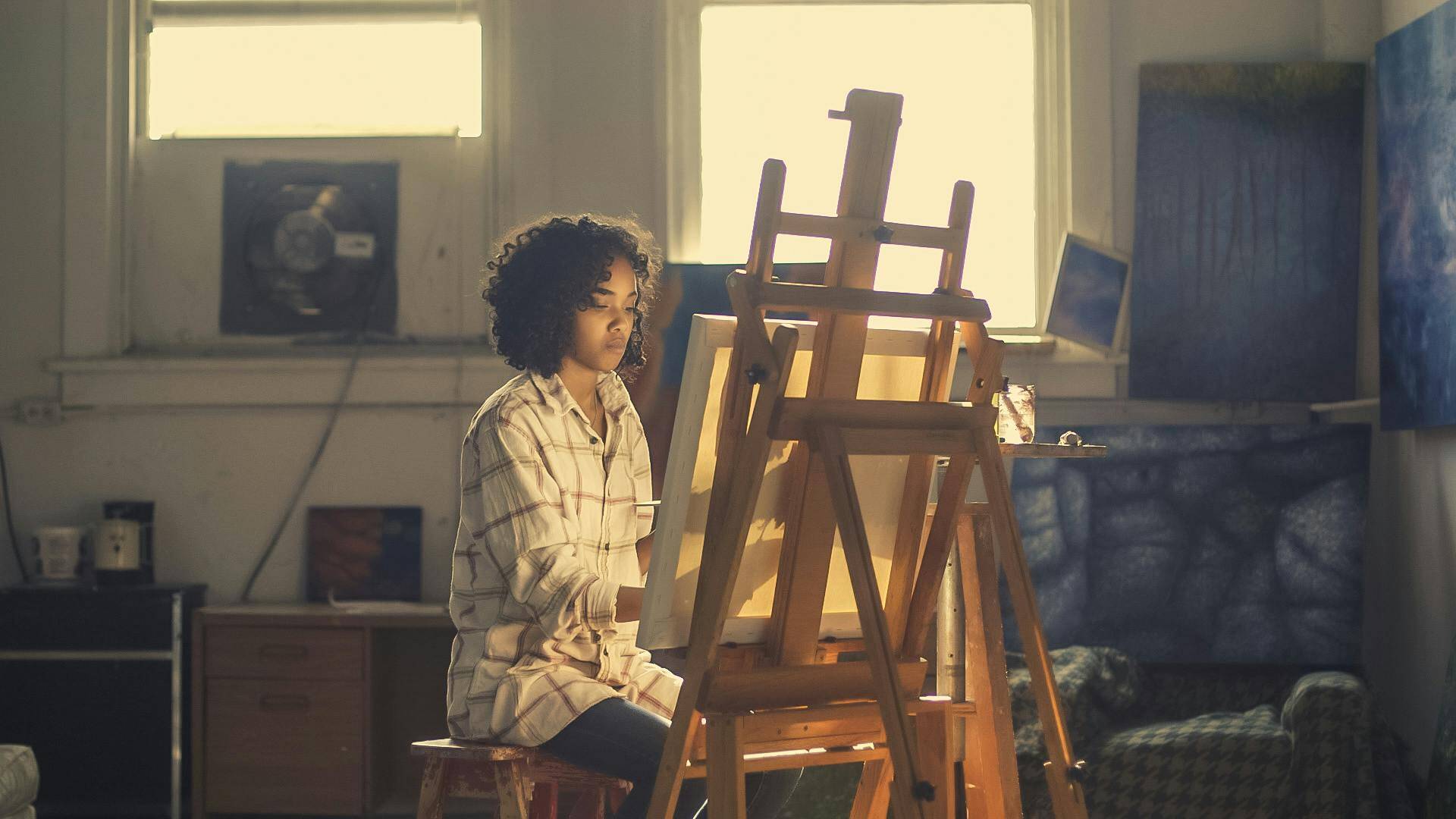 A young woman pursues her passion as she sits quietly painting on a large canvas in a artist's studio