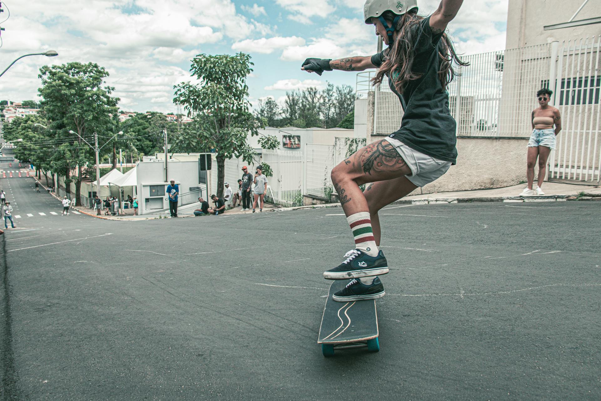  A young woman is skateboarding down a steep, urban street. She is wearing a helmet, black t-shirt, white shorts, and knee-high striped socks. Her arms are outstretched for balance as she maneuvers the skateboard, showcasing her tattoos on her legs. The background features several onlookers, a mix of people standing and sitting by the roadside. 